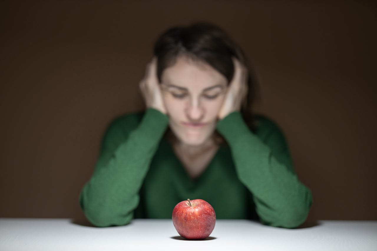 Home A woman in a green sweater observes an apple on a table, creating a thoughtful scene.